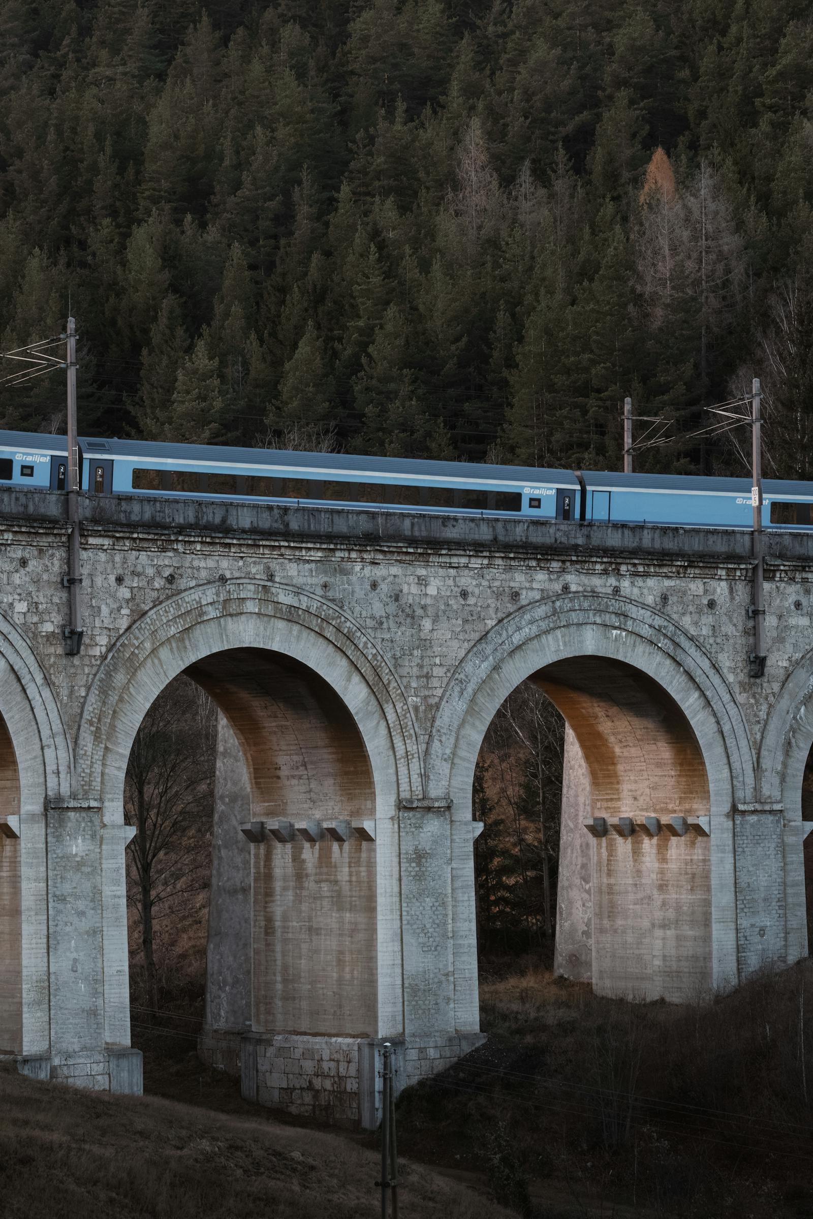 A blue train travels over the historic Semmering Railway viaduct amidst a lush forest in Austria.