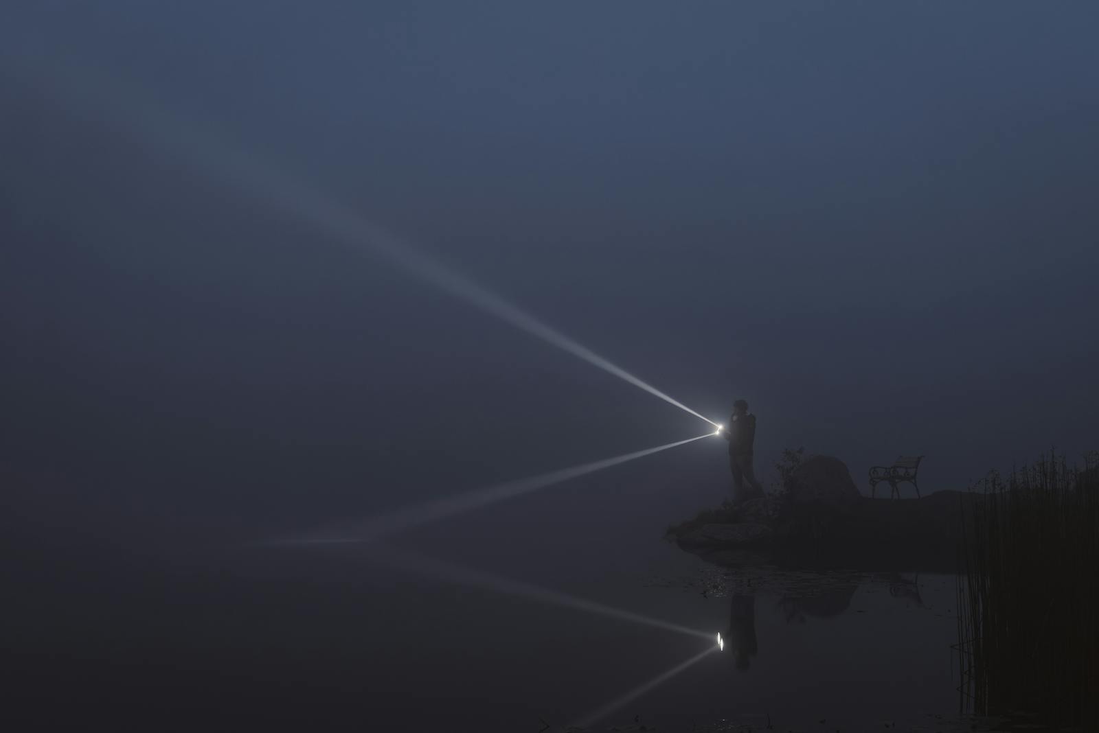 A solitary figure illuminates a foggy lakeside at night with a flashlight.