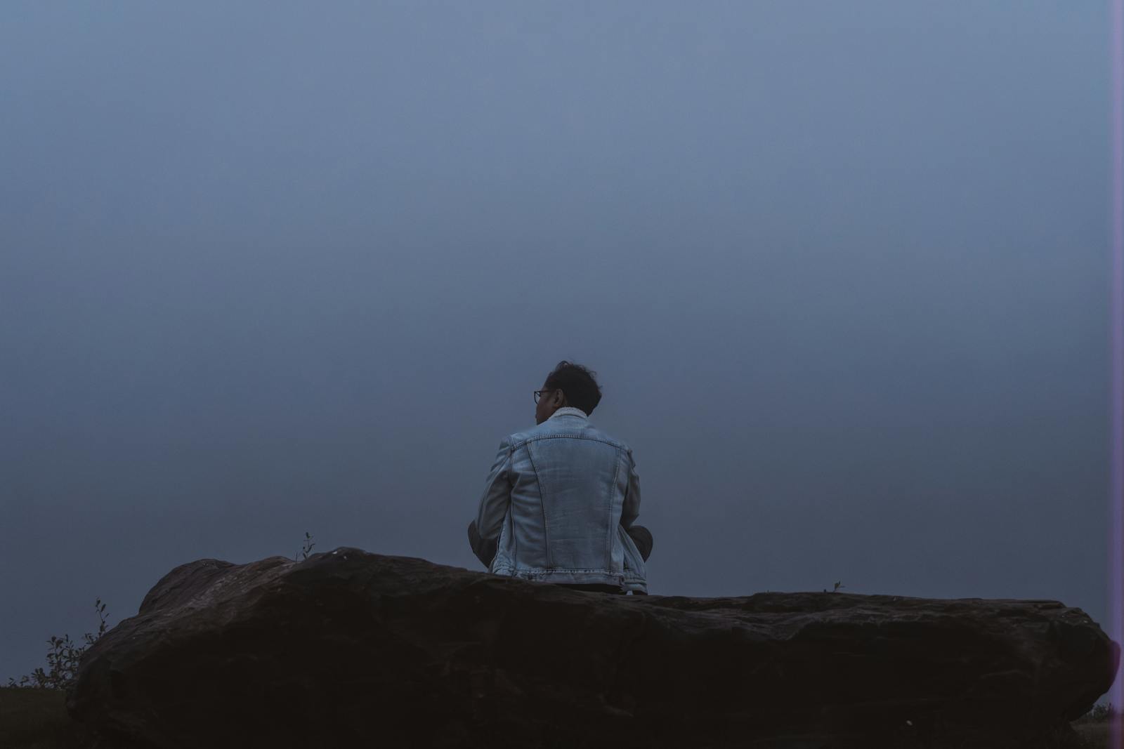 A person sitting on a rock in a foggy evening setting, evoking introspection and solitude.