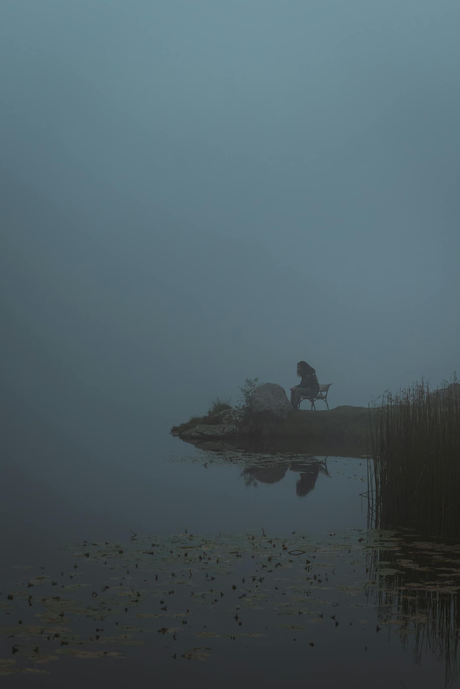 Silhouette of a person sitting by a misty lake, reflecting in still water.