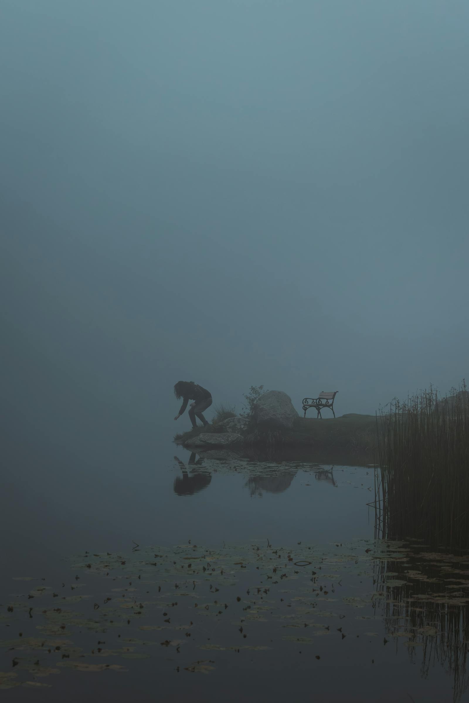 Shadowy figure jumping by a foggy lake with a bench and reeds, Styria, Austria.