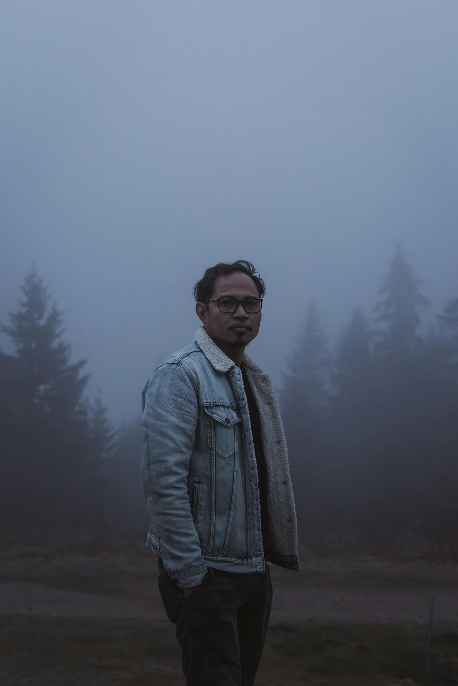 A man in a denim jacket stands amidst a foggy forest in Styria, Austria.