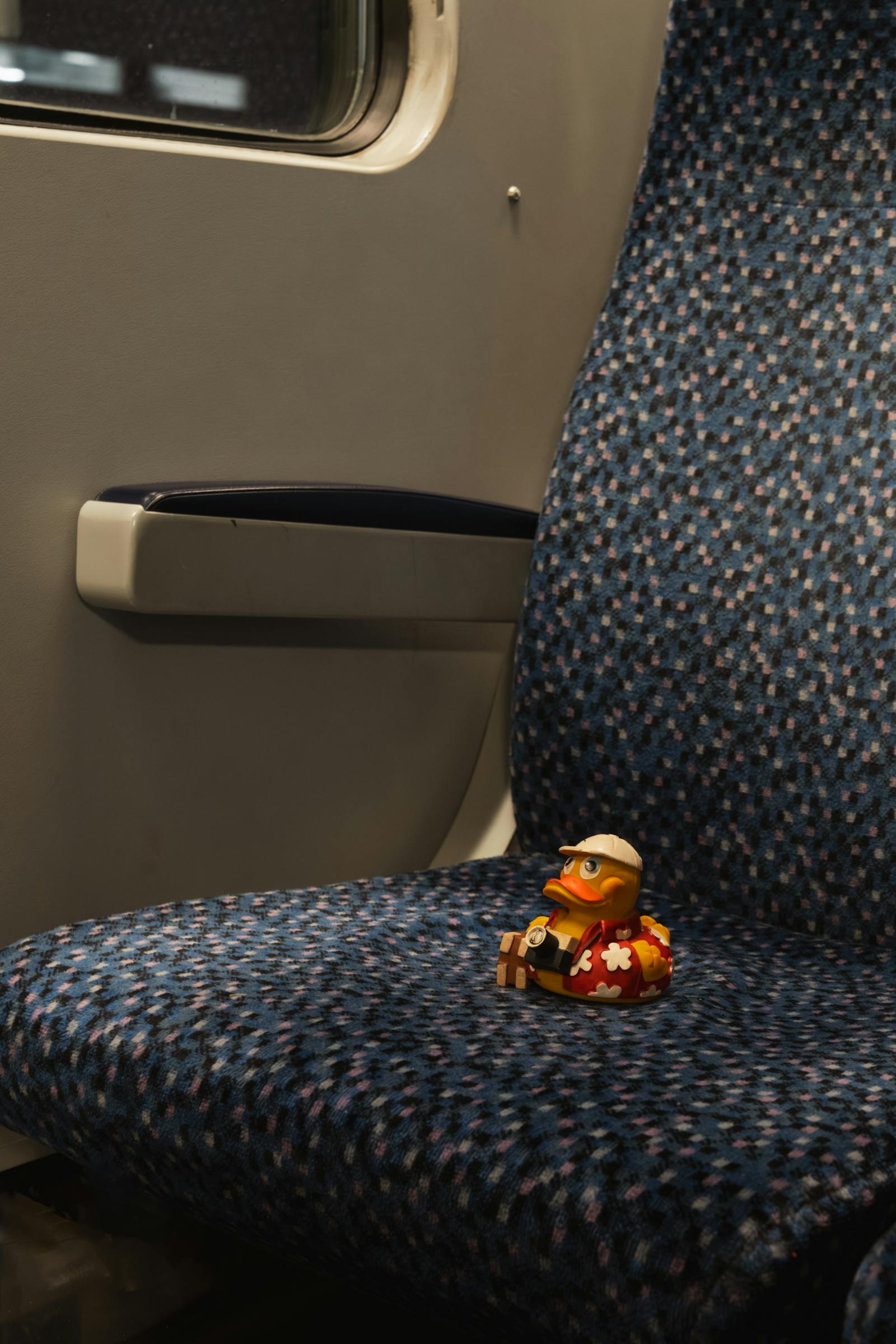 A small toy duck with a quirky design sits alone on a train seat, symbolizing unexpected travel adventures.