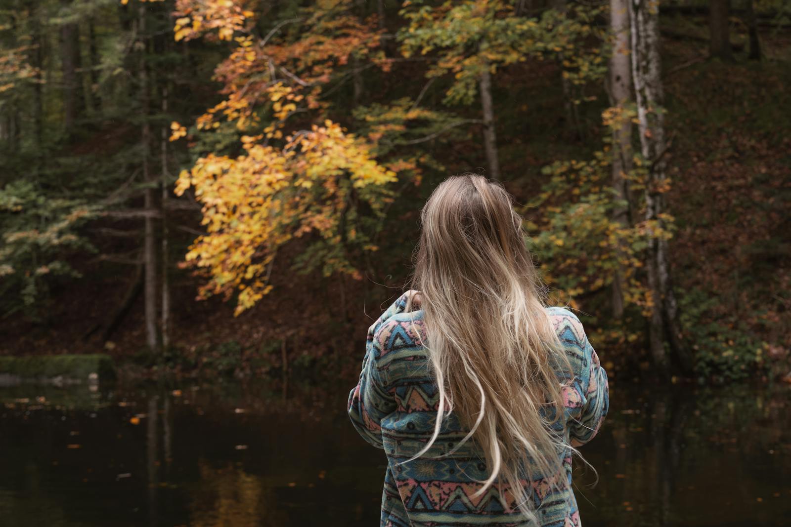 A woman in a sweater admires the vibrant autumn foliage by a tranquil lake in a serene forest setting.