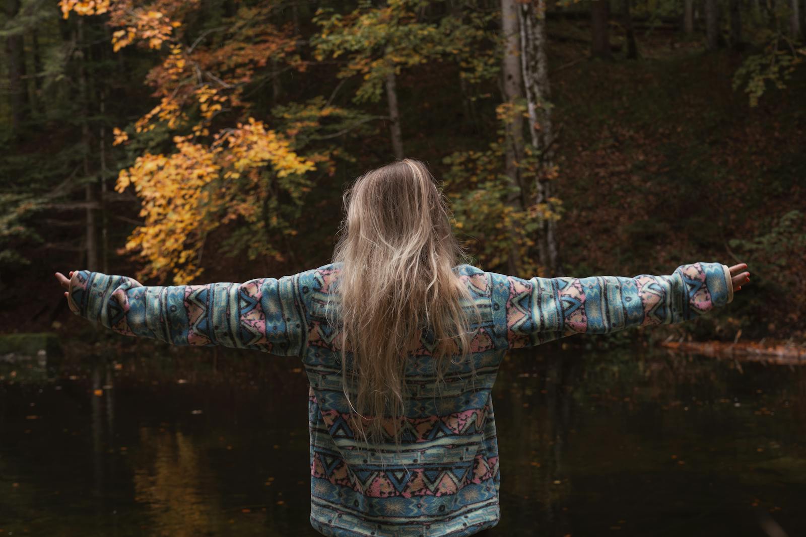 Back view of woman embracing nature with open arms in forest during fall.