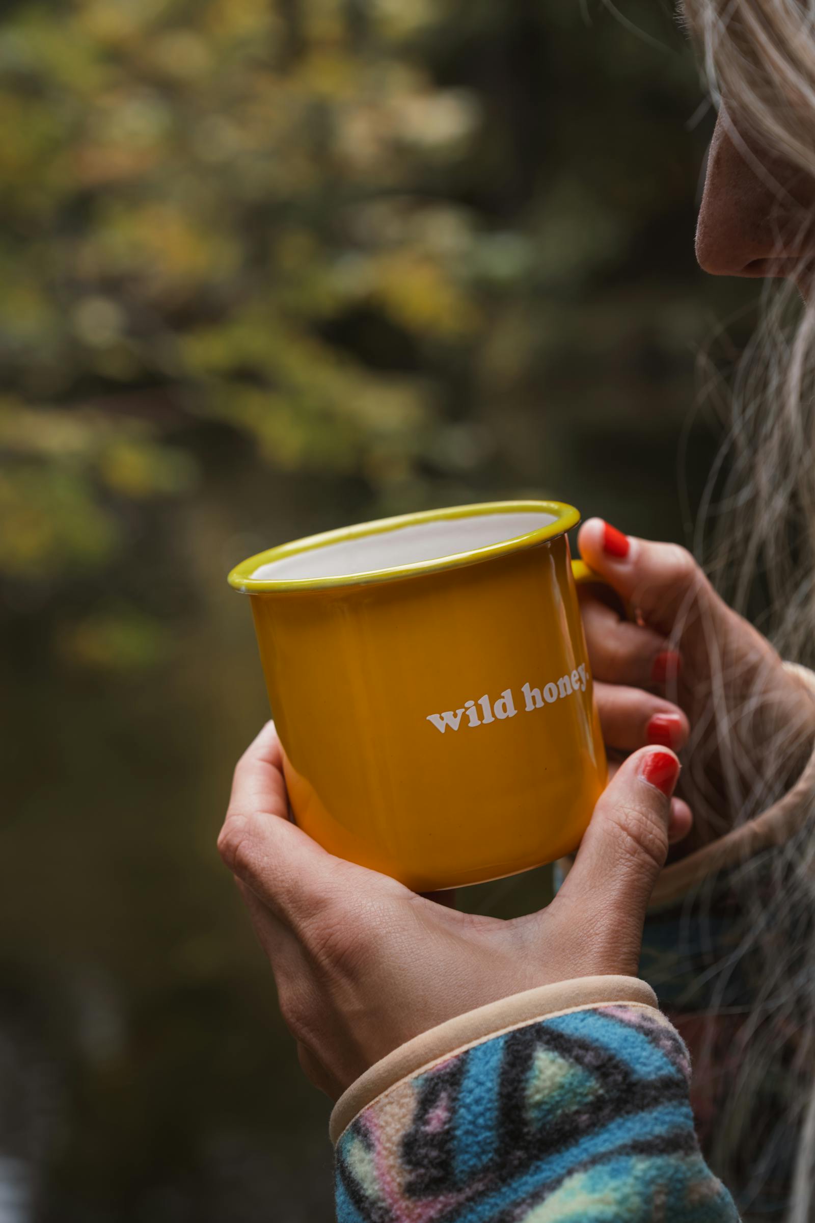 Woman holding yellow mug marked 'wild honey' amidst fall foliage, evoking cozy vibes.