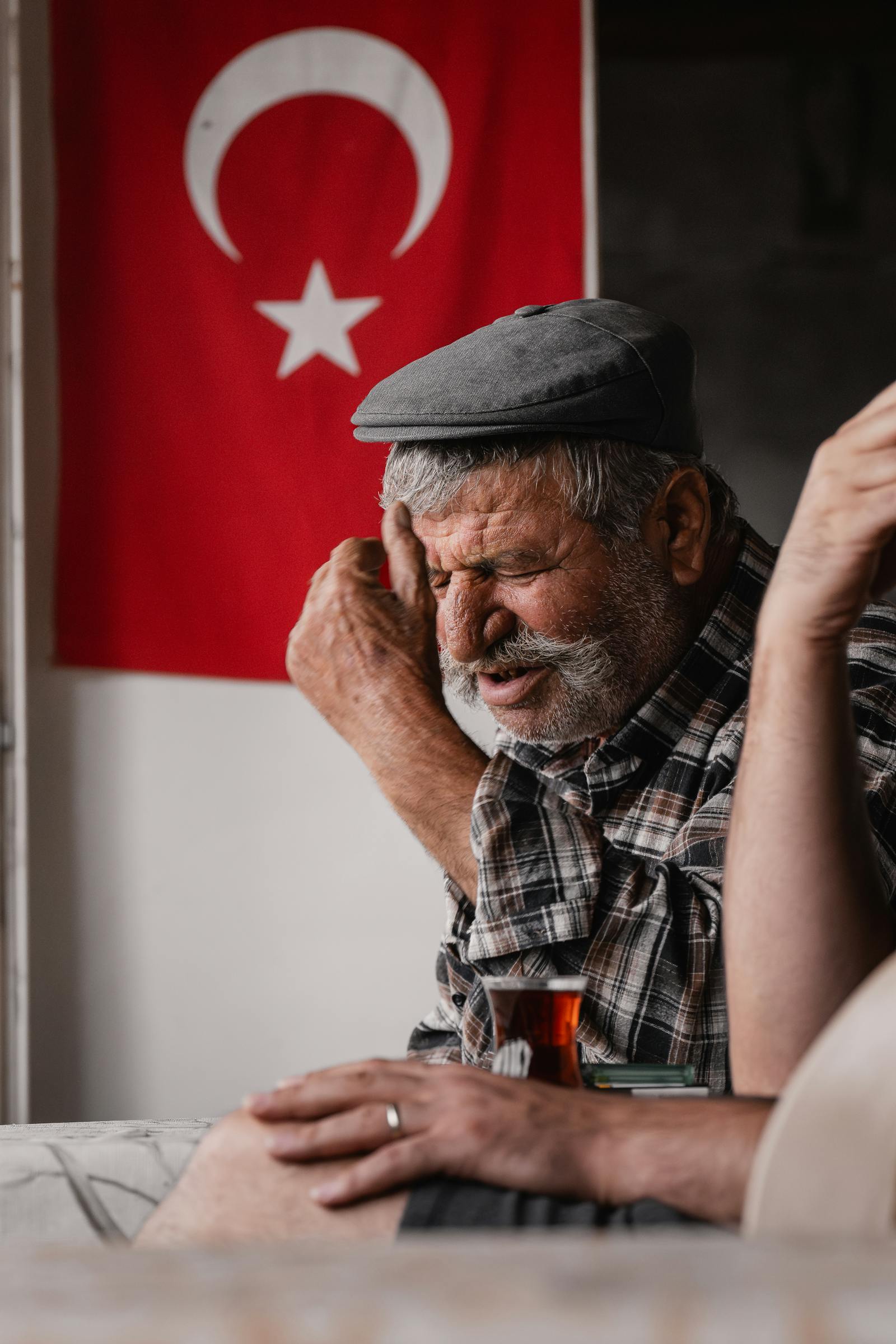 Portrait of an elderly man in Turkey enjoying tea and conversation indoors, with a Turkish flag in the background.