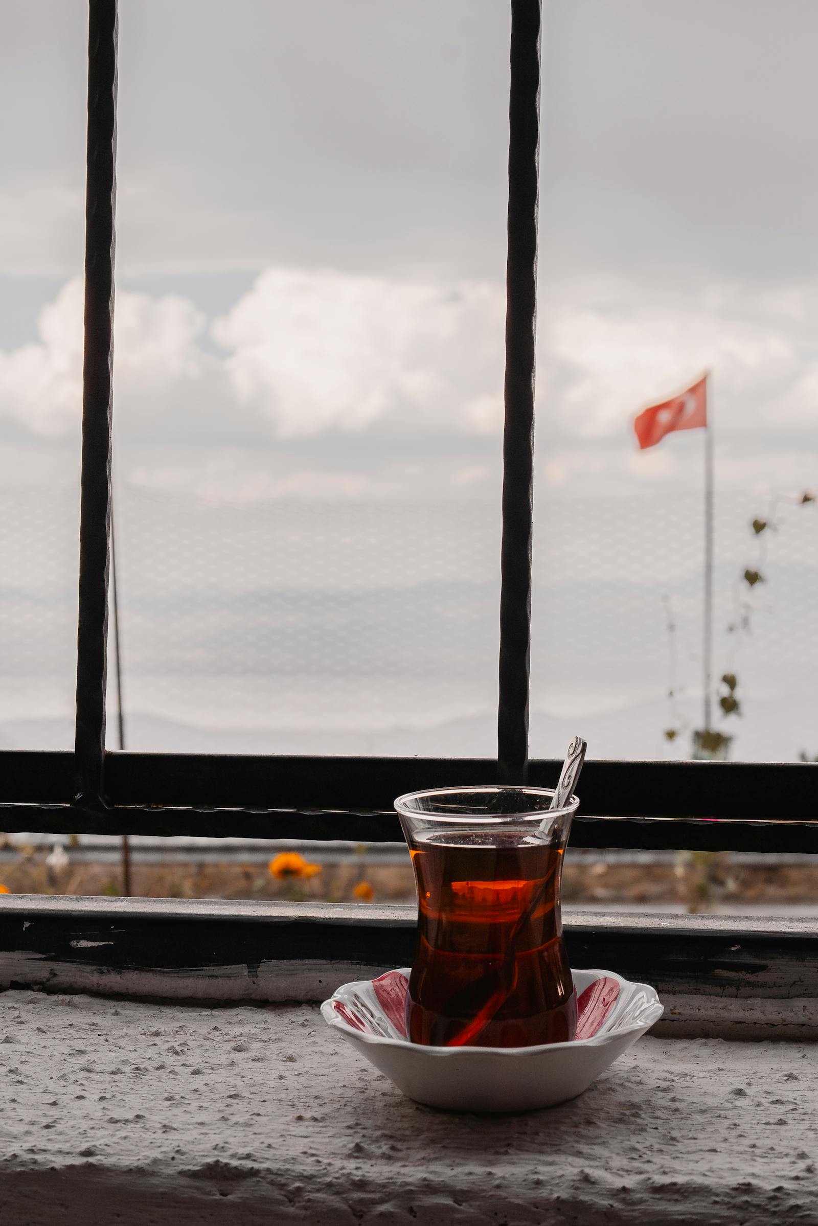 A glass of Turkish tea on a windowsill with a scenic view of clouds and a Turkish flag outdoors.