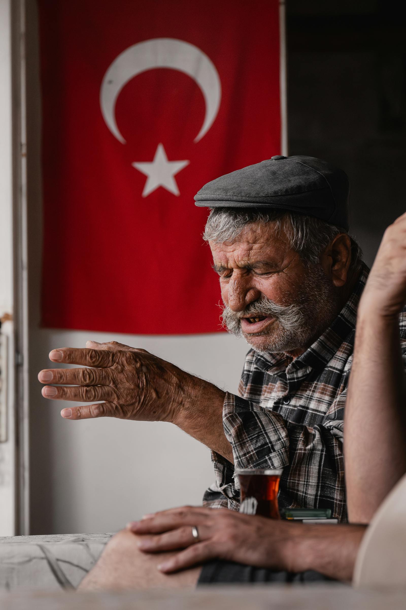 Elderly man engrossed in storytelling with a Turkish flag in the background, indoors.