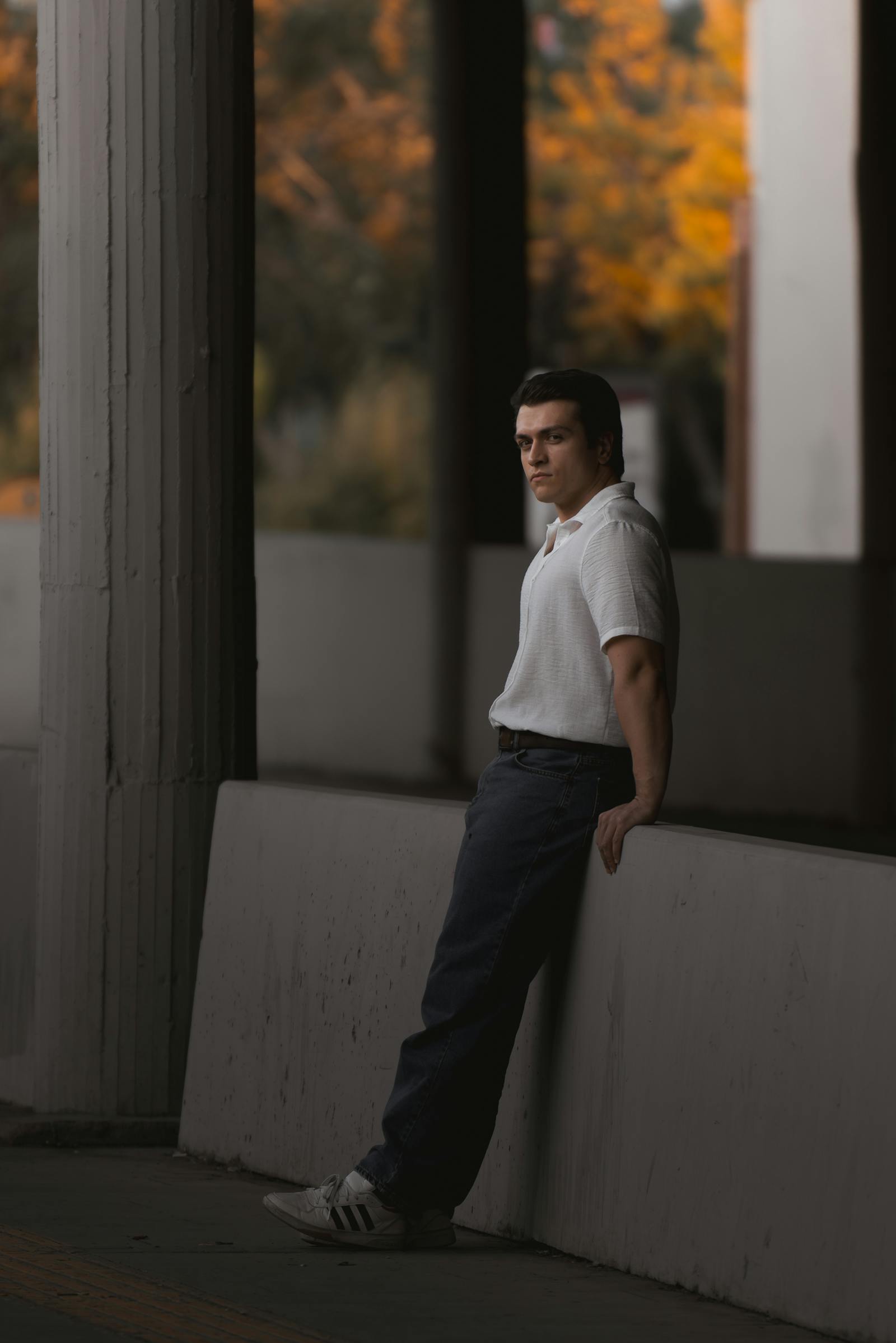 A thoughtful man leans on a wall in an urban area, with autumn colors in the background.