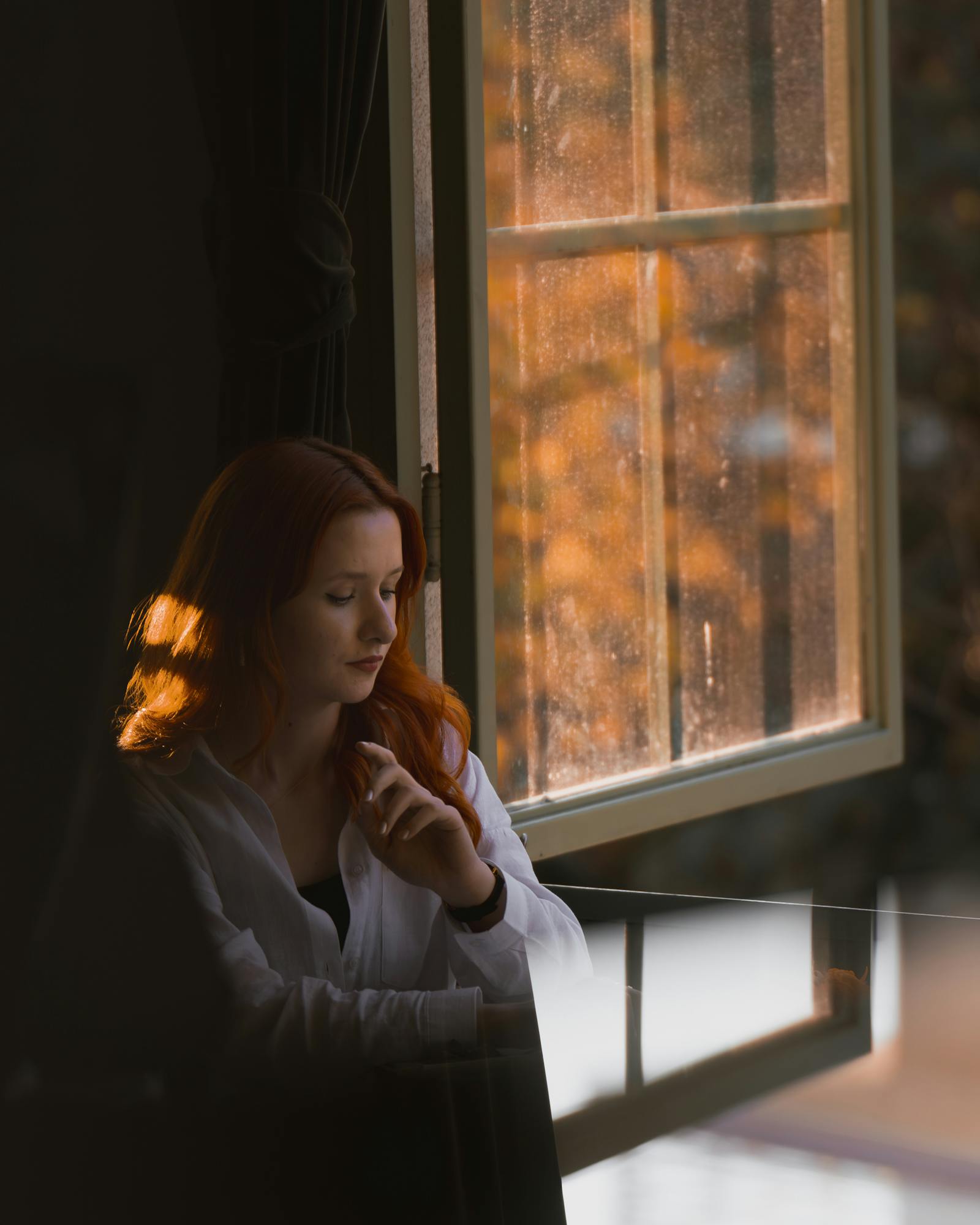 A woman with red hair gazes pensively out a sunlit window in a cozy room.