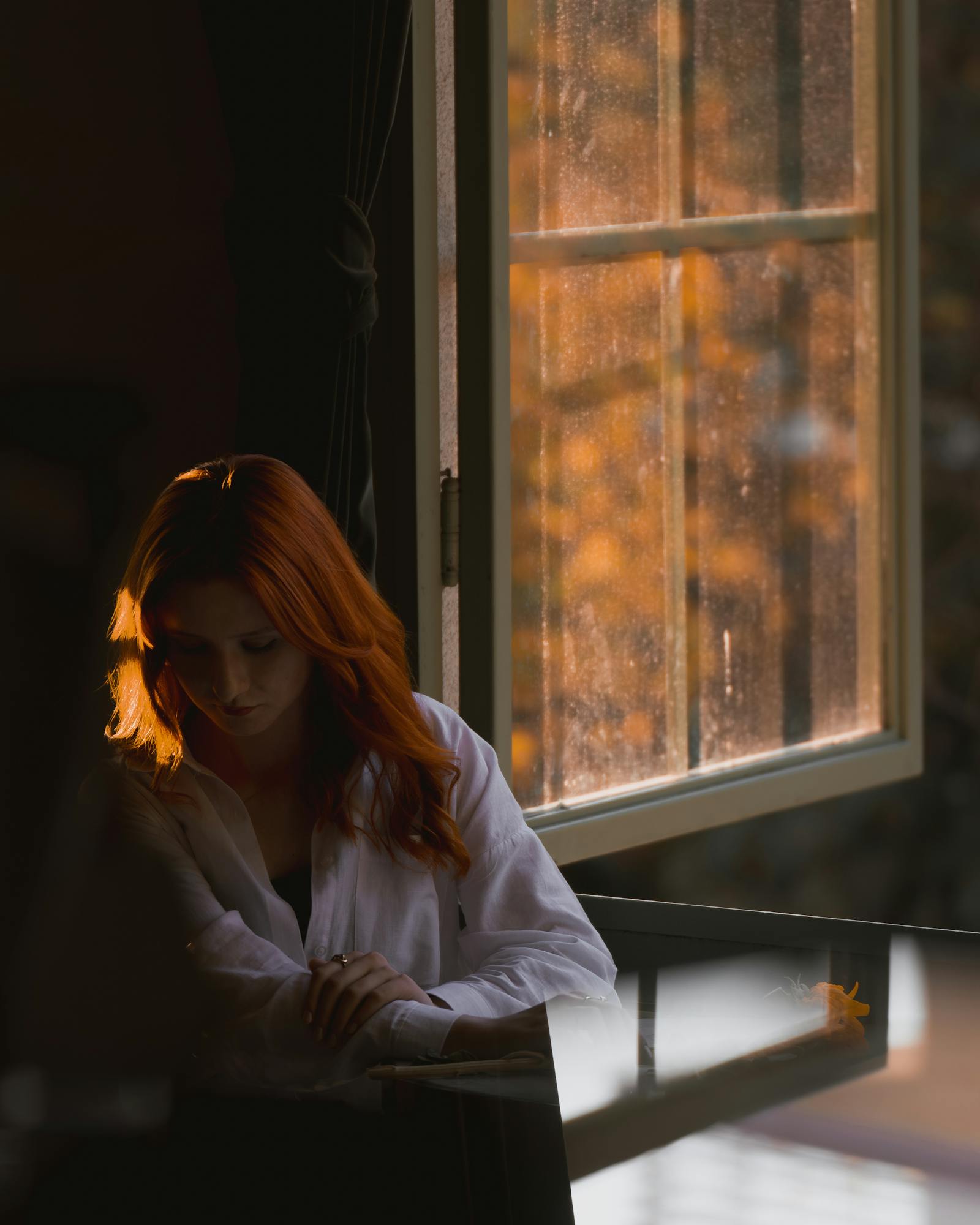 Introspective woman with red hair sits by a window during golden hour in fall.