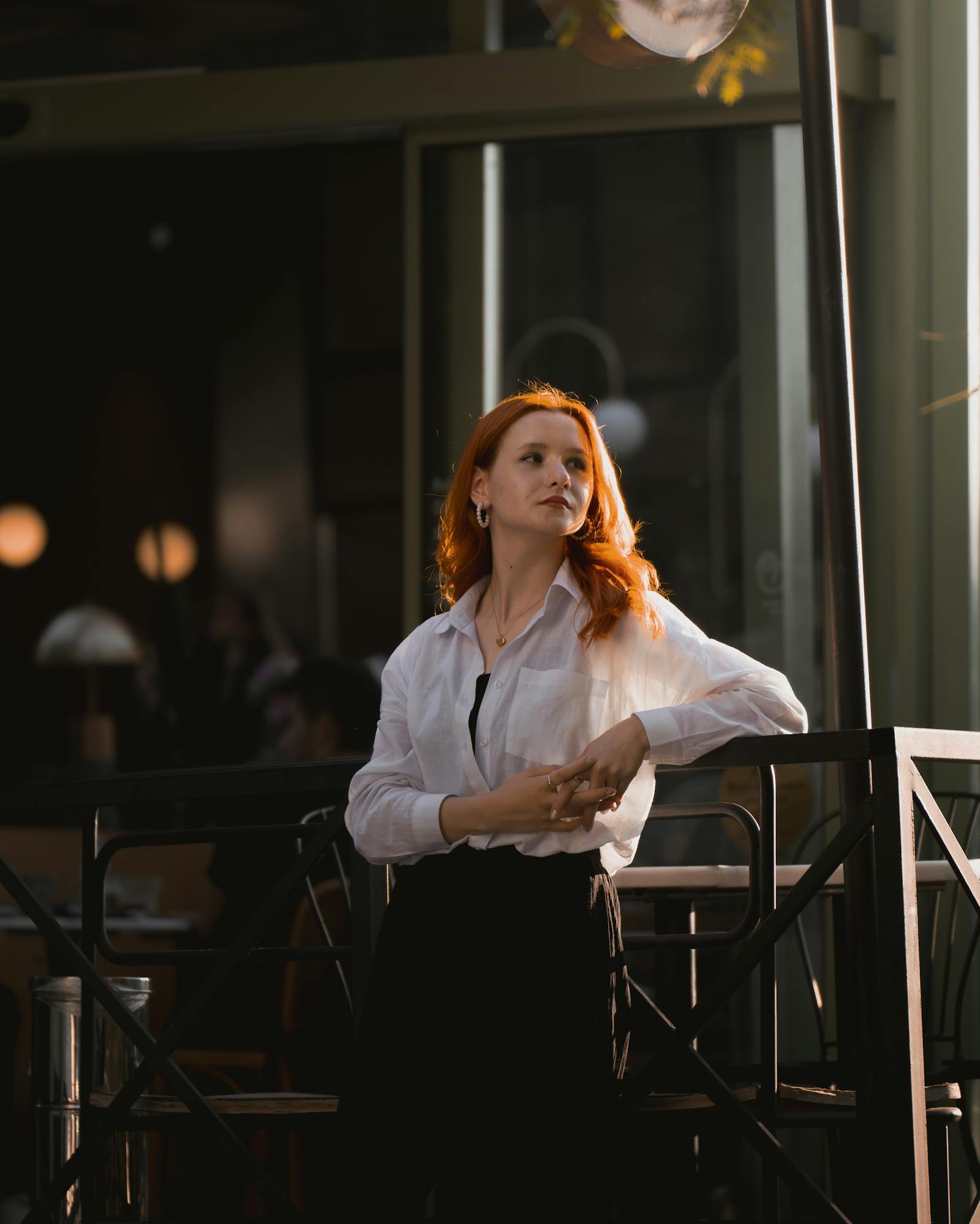 Elegant woman in white blouse with red hair poses at a chic café, dramatic lighting highlights her confident demeanor.