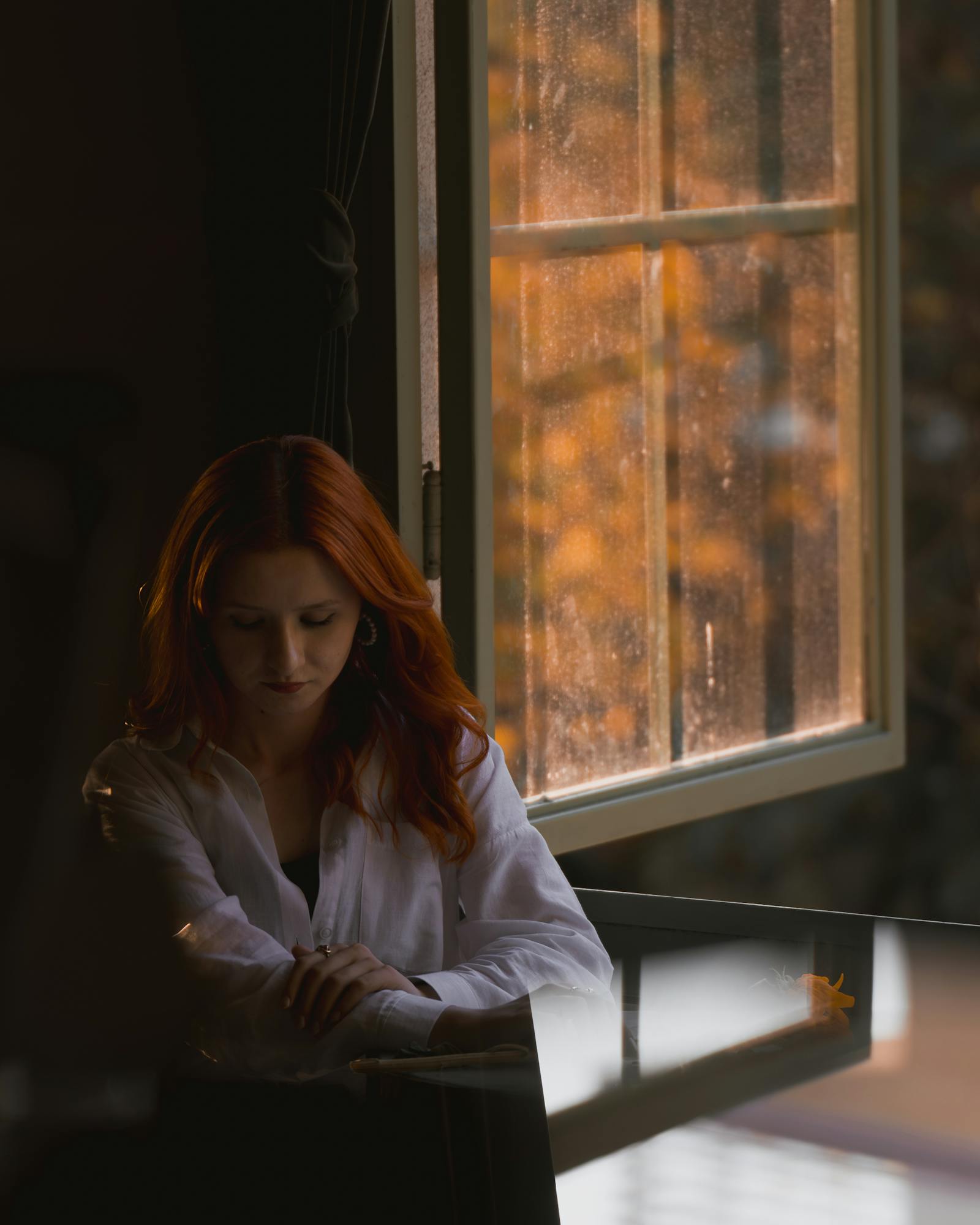 A red-haired woman sits introspectively by an open window with autumn leaves outside.