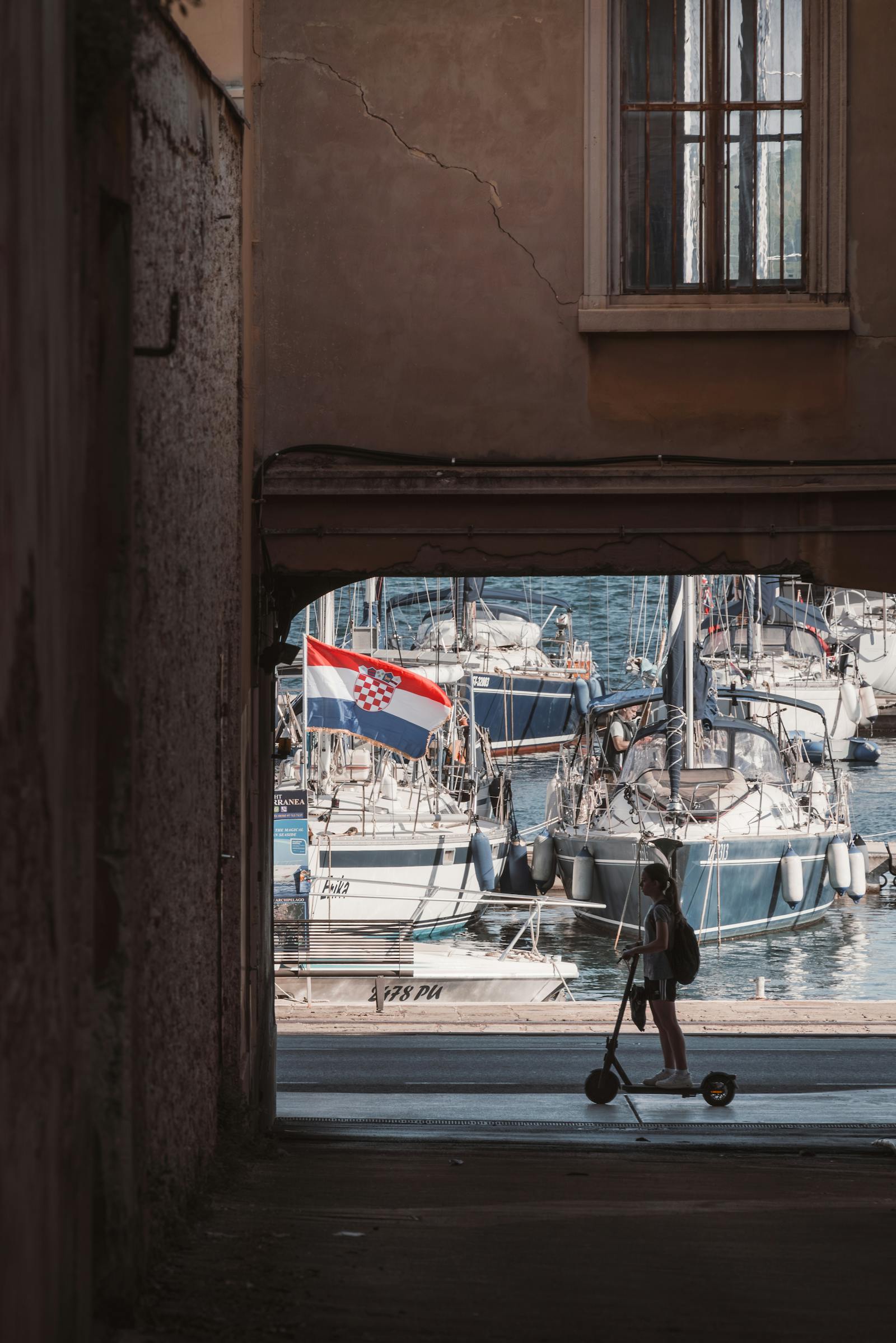 Silhouette of a person riding an electric scooter by the harbor in Pula, Croatia, with moored yachts and Croatian flag.