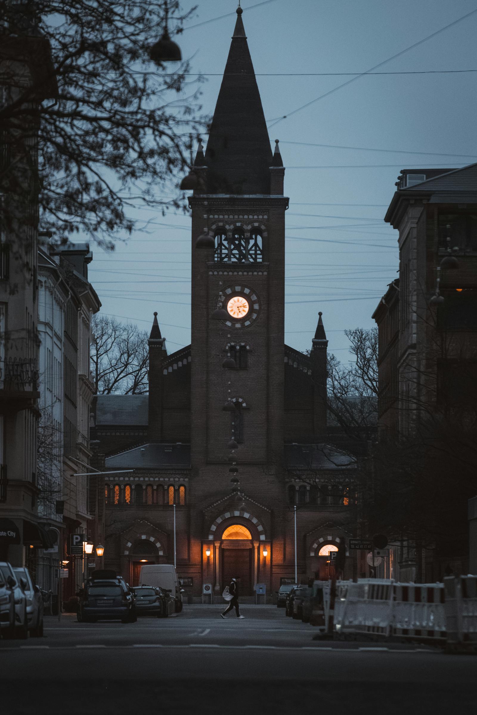 A gothic church tower with a glowing clock face at dusk in an urban street view.