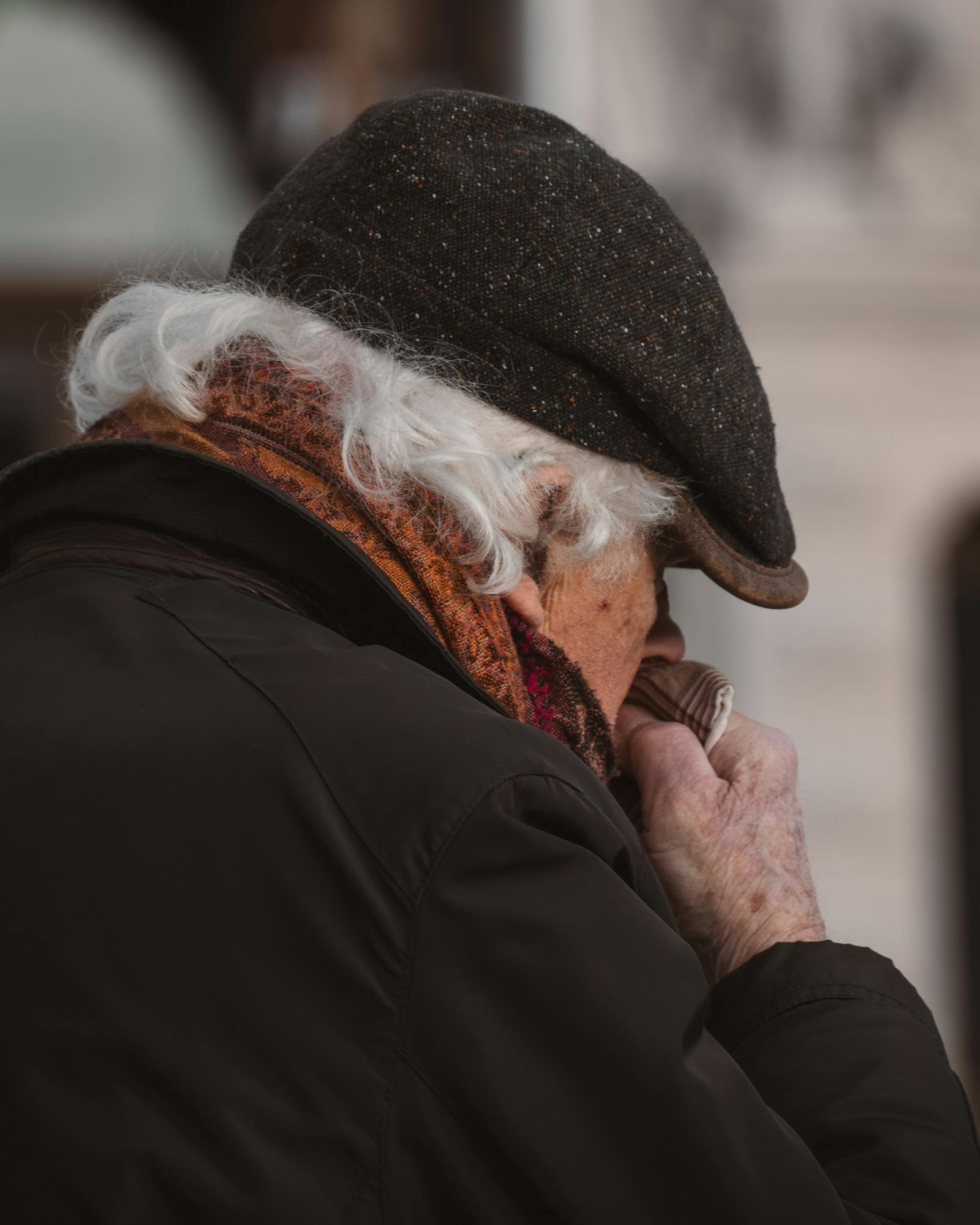 Senior man with gray hair wearing an ivy cap and scarf, blowing nose on a winter day in Stockholm.