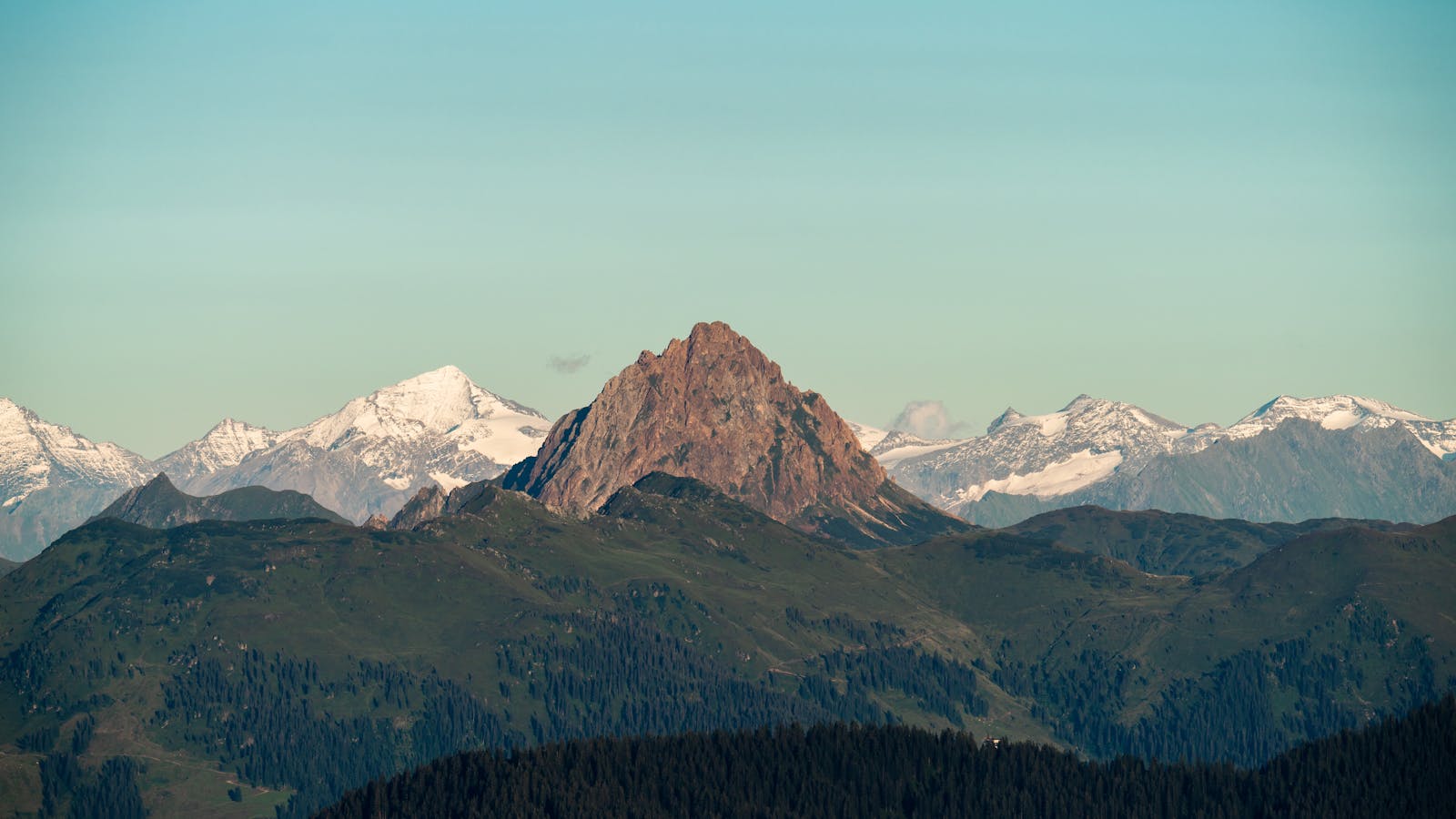 Breathtaking panoramic landscape of snow-capped mountains and lush green hills under a clear sky.