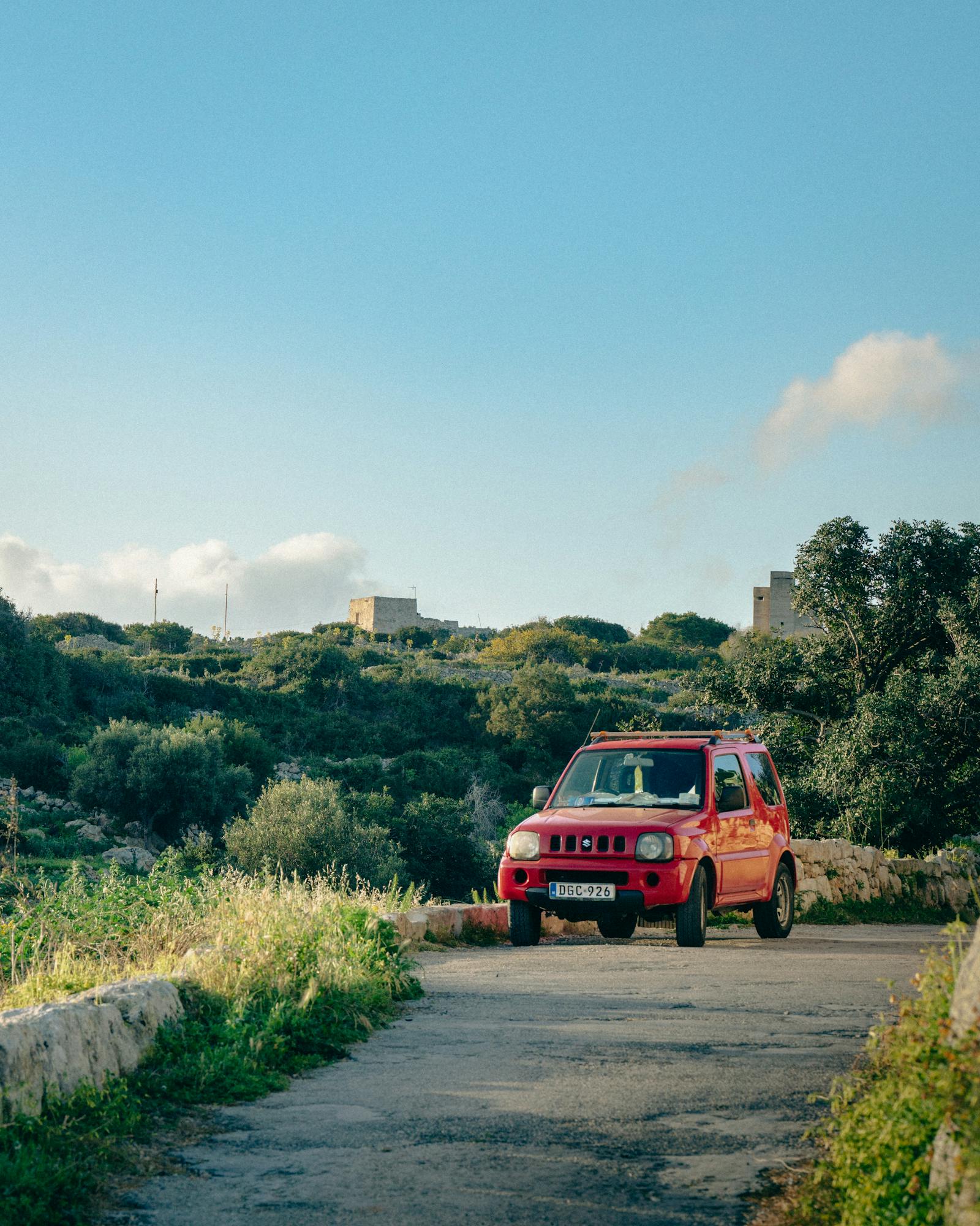 Red SUV driving on a rural road with lush countryside and clear blue sky.