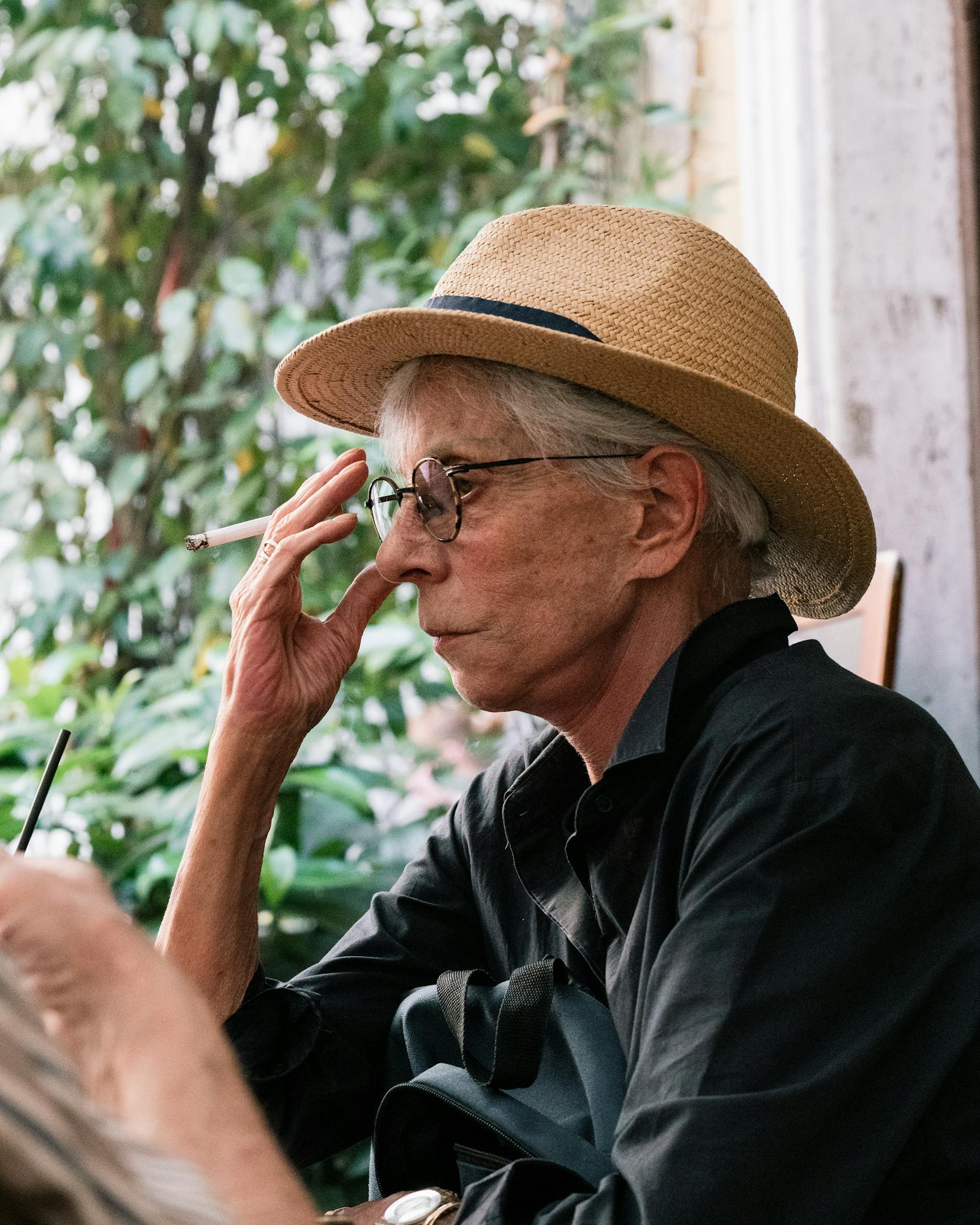 Elderly woman in a hat sits outdoors, smoking a cigarette in Rome, Italy.