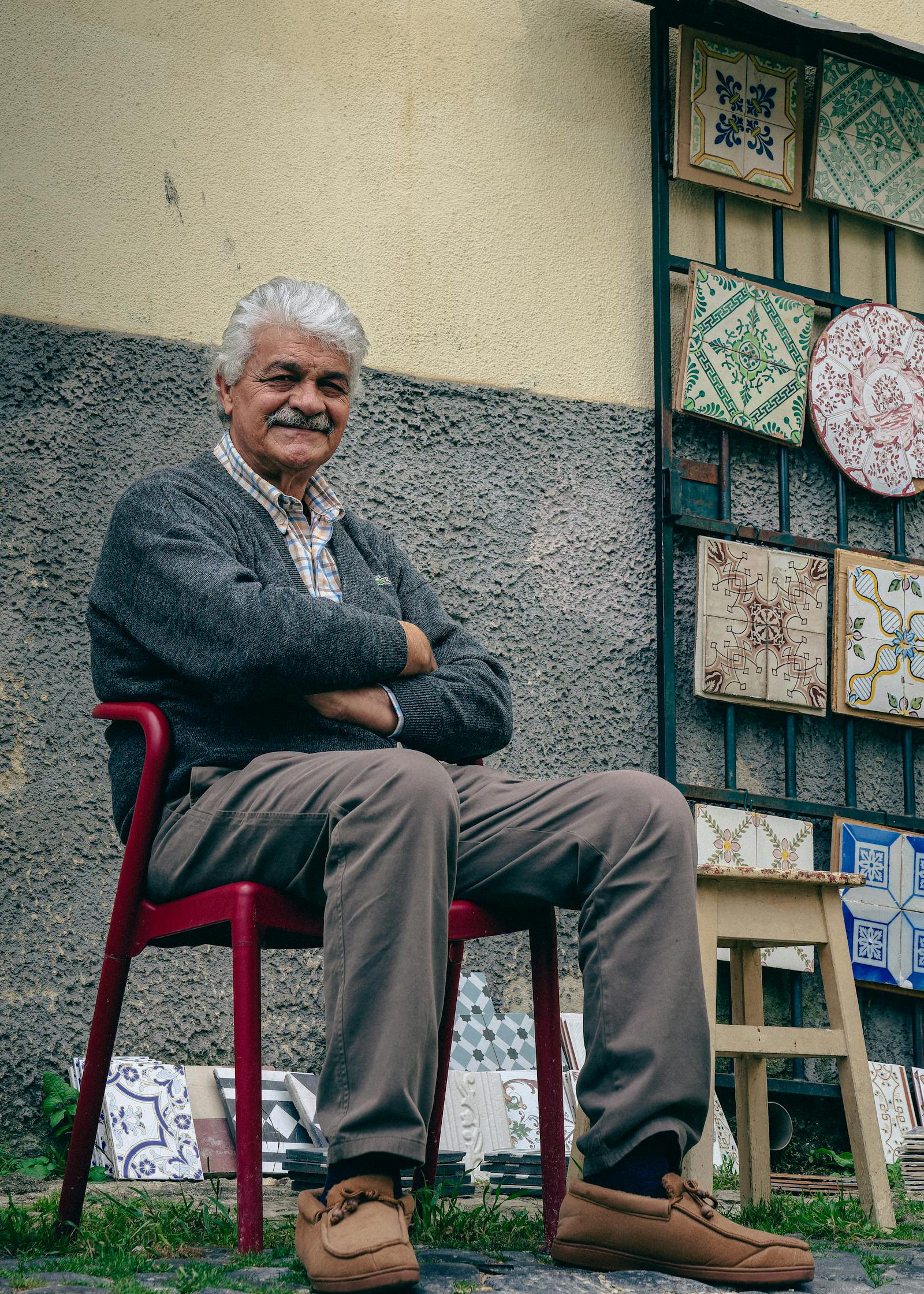 Elderly man with crossed arms sits by colorful tiles in Lisbon street.