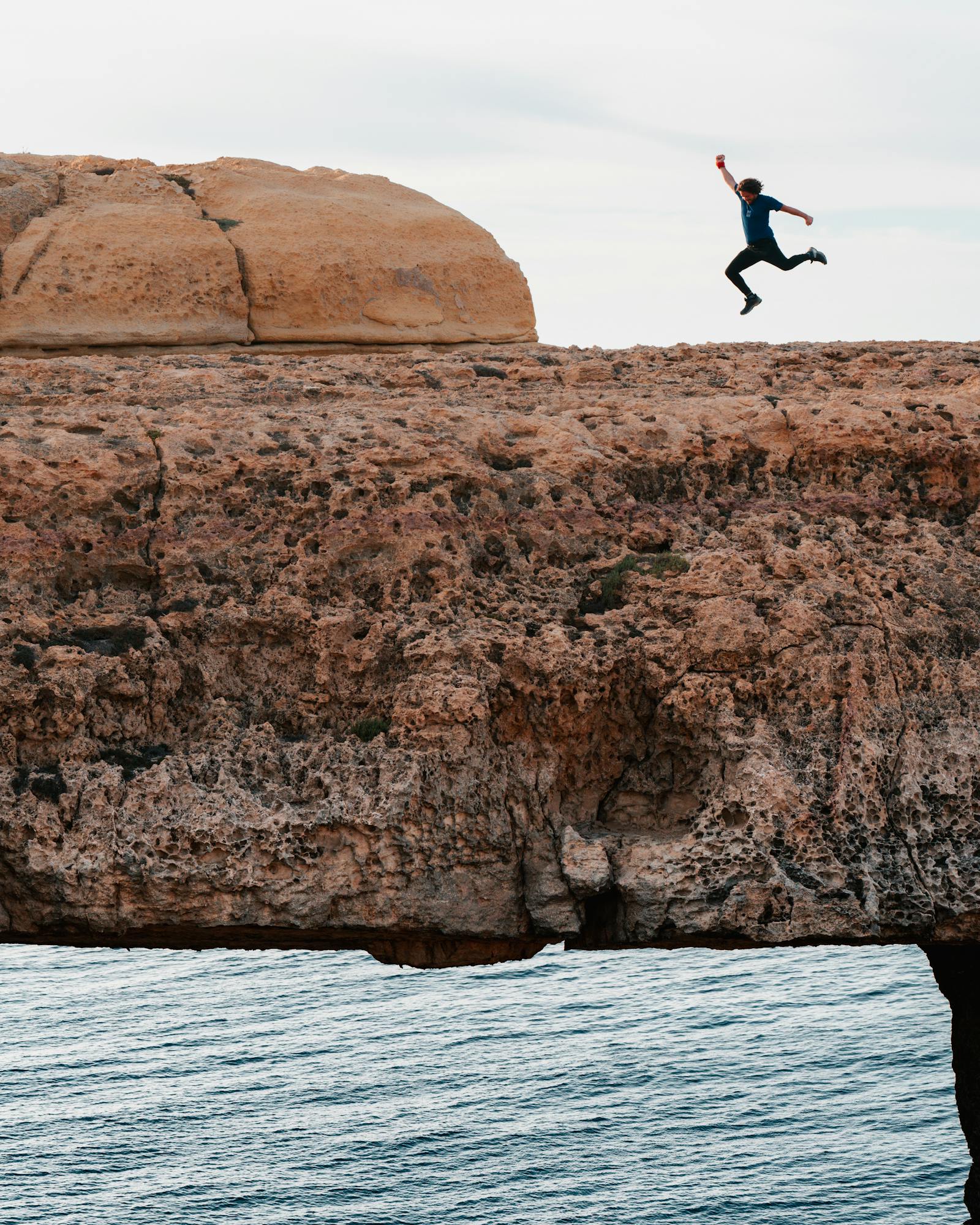 Man leaps across rocky arch over ocean, showcasing thrill and adventure in Malta.